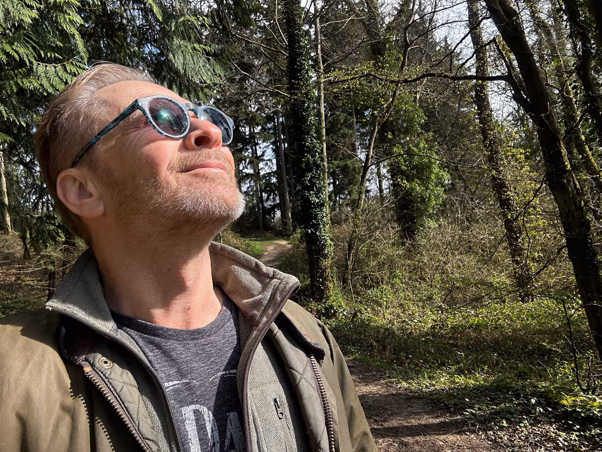 A man wearing sunglasses looks up while standing in a wooded area.