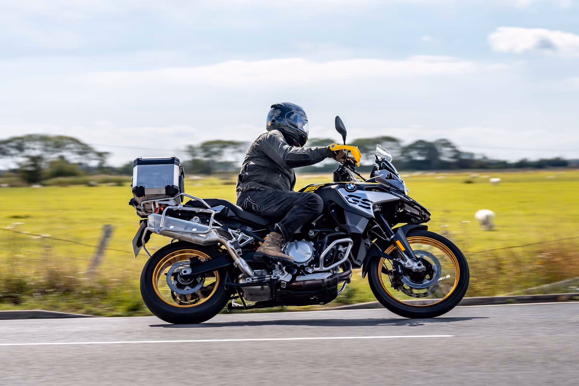 Person riding a motorcycle on a rural road with fields and sheep in the background.