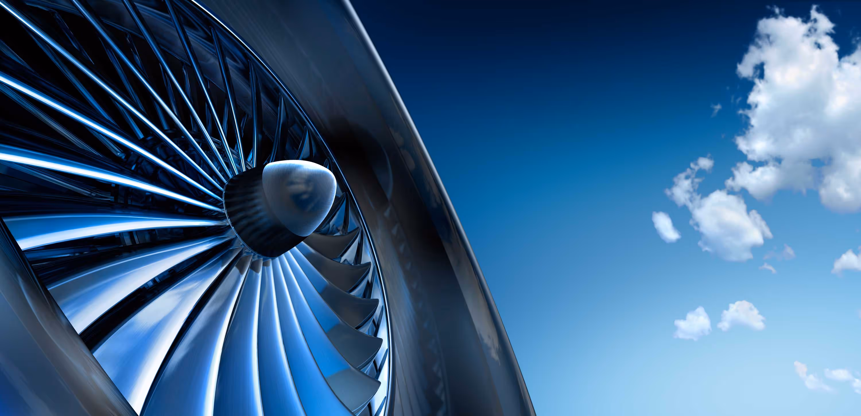 Close-up view of an aircraft jet engine turbine, with a clear blue sky and clouds in the background.
