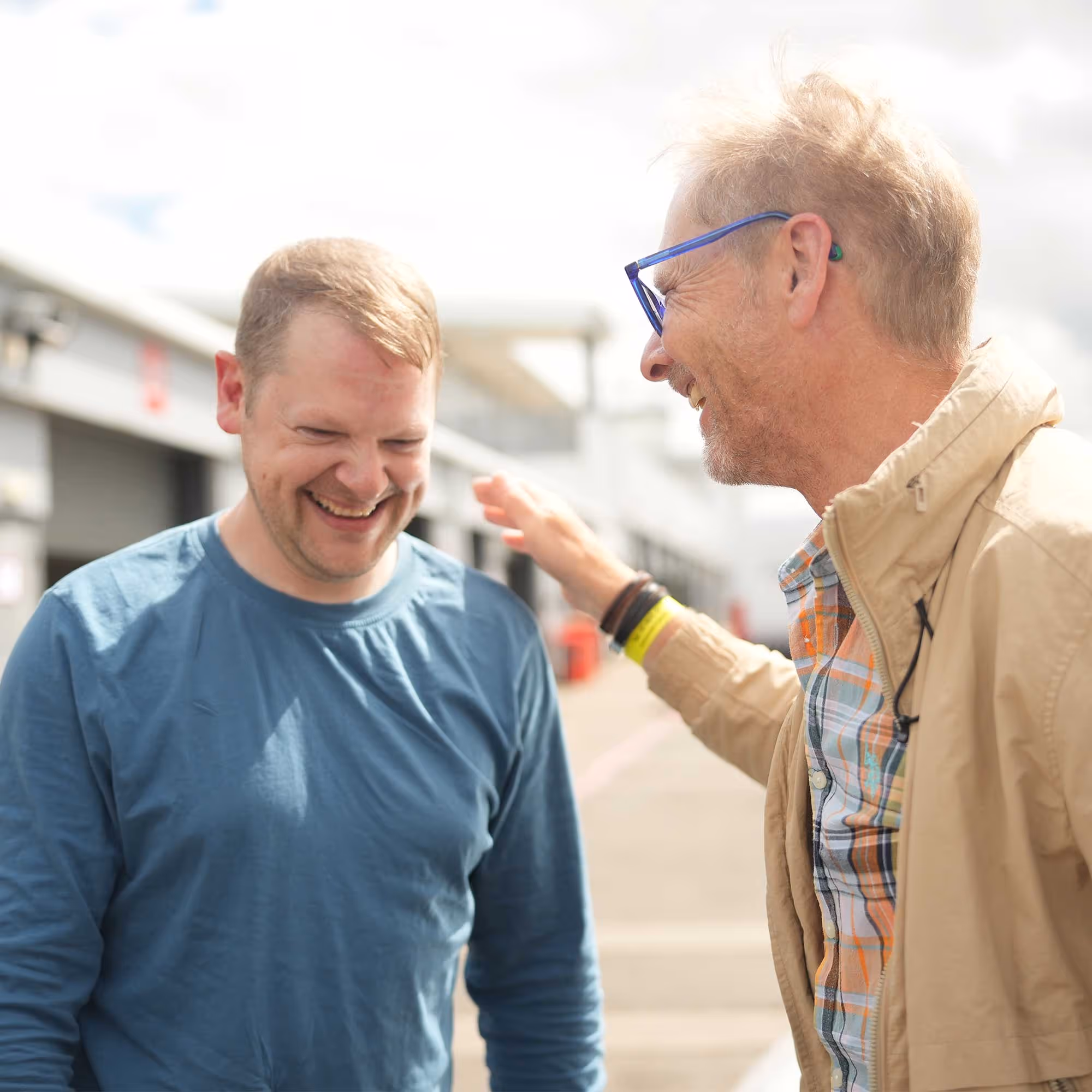 Two men standing, smiling, one pats the other's shoulder, outside near a building.