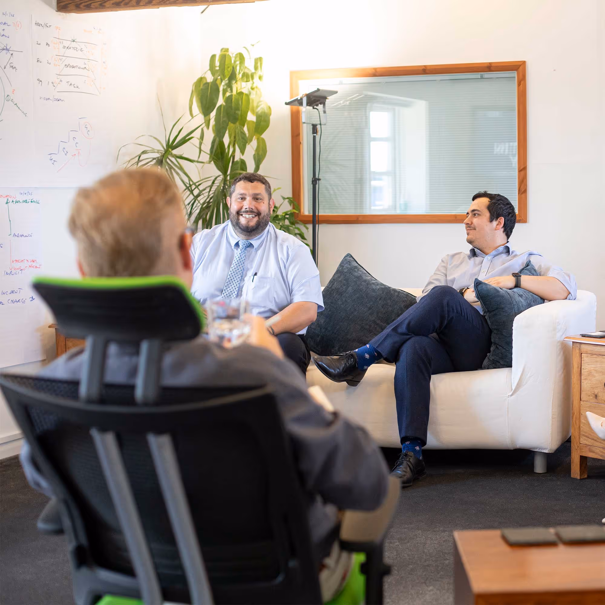 Three men sitting in an office having a discussion, with one facing away in a chair.