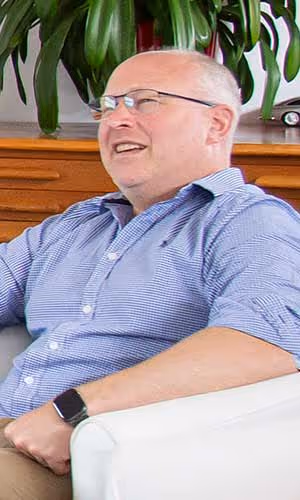 A man sitting and smiling, wearing glasses and a blue shirt.