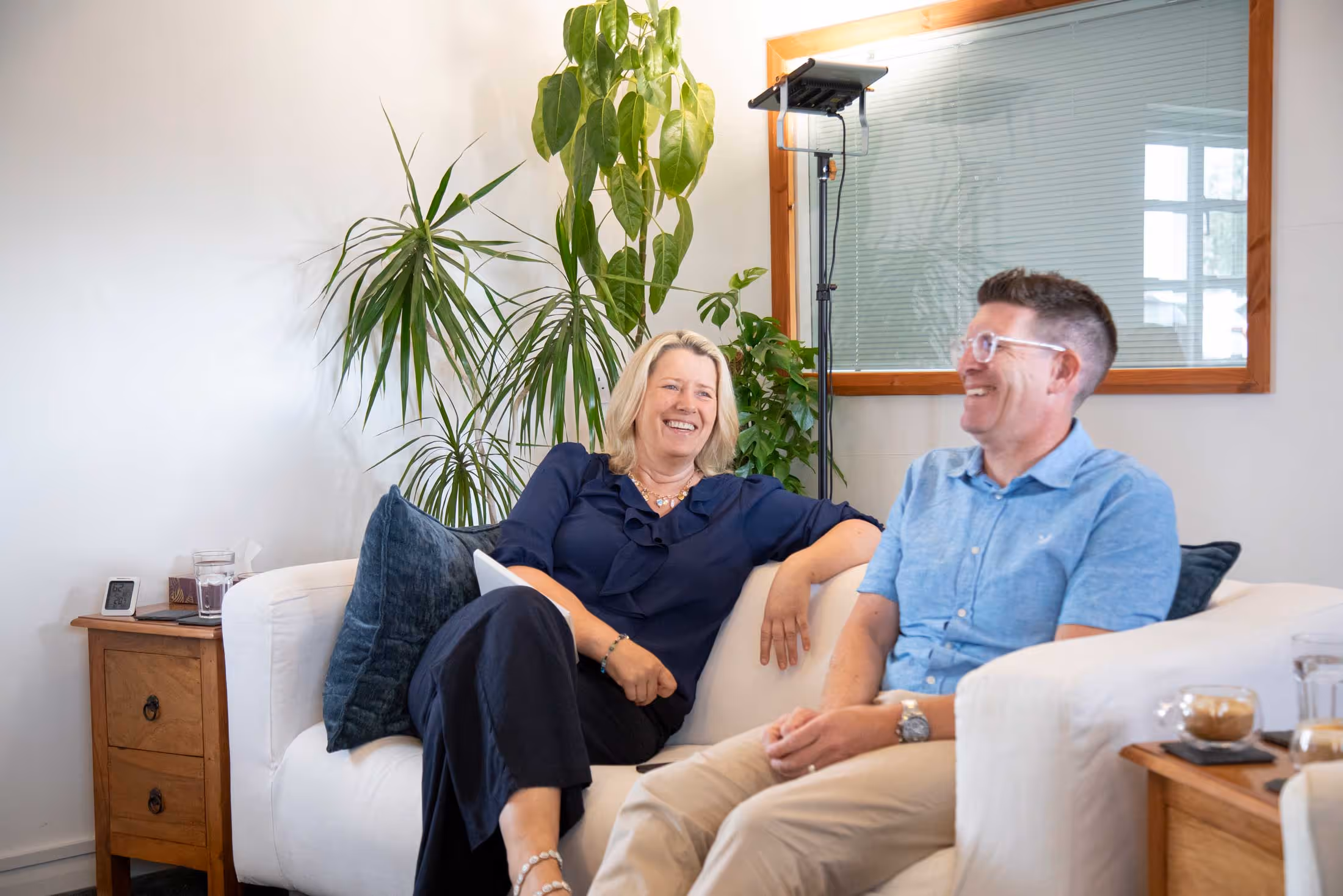 Two people sitting on a sofa, smiling and talking, next to indoor plants and a lamp.