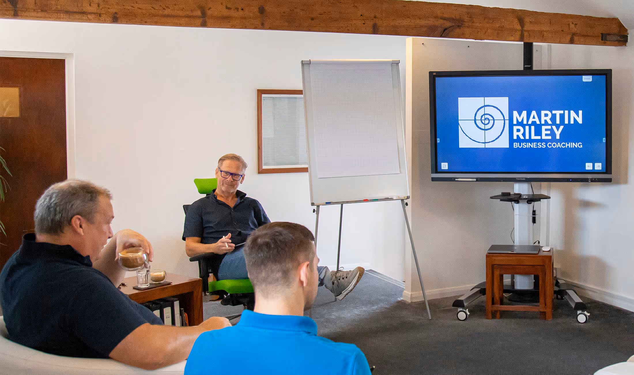 Three men in a meeting room watching a business coaching presentation on a large screen.