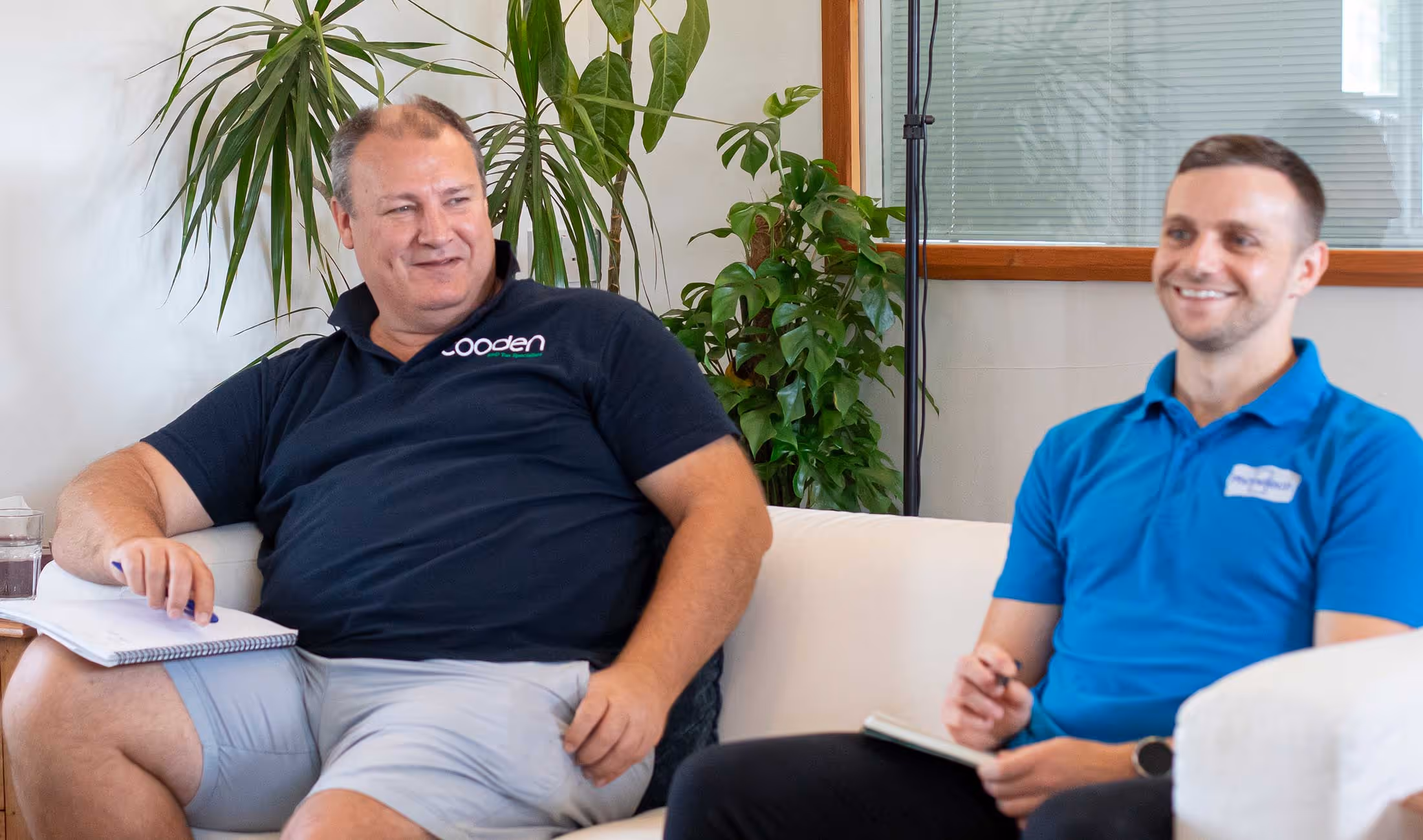 Two men sitting on a couch, smiling, and holding notebooks during a casual meeting.