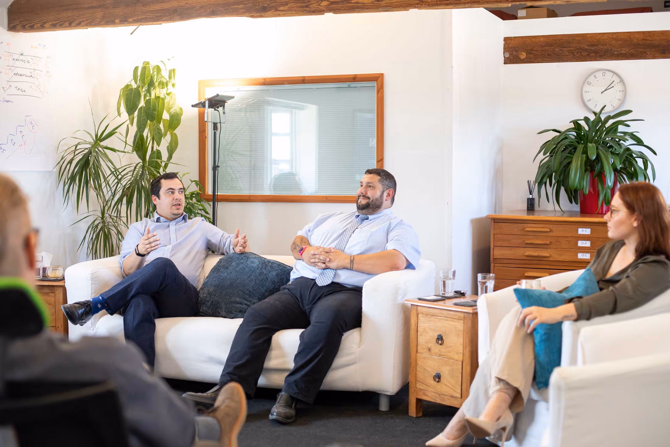 Three people sitting and discussing in a bright office with plants and a clock on the wall.