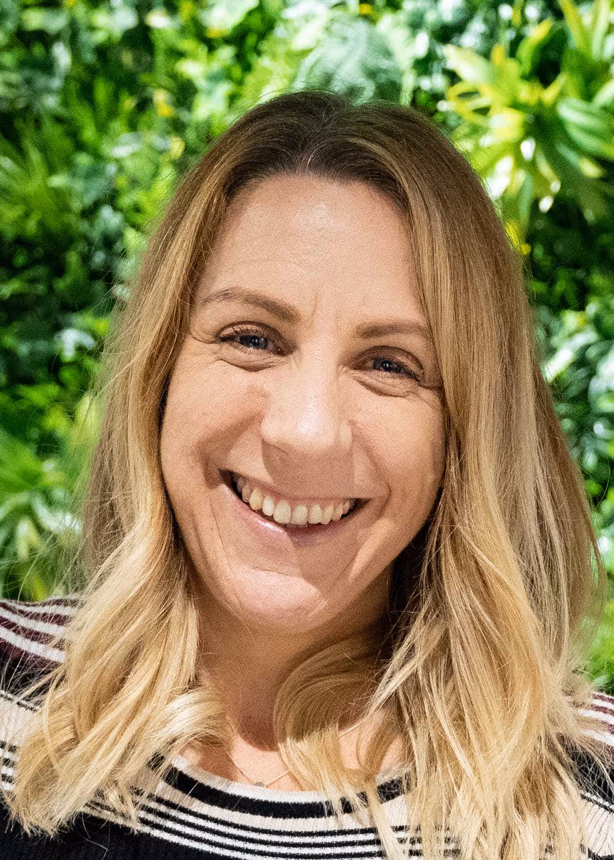A person smiling in front of a wall filled with green plants.