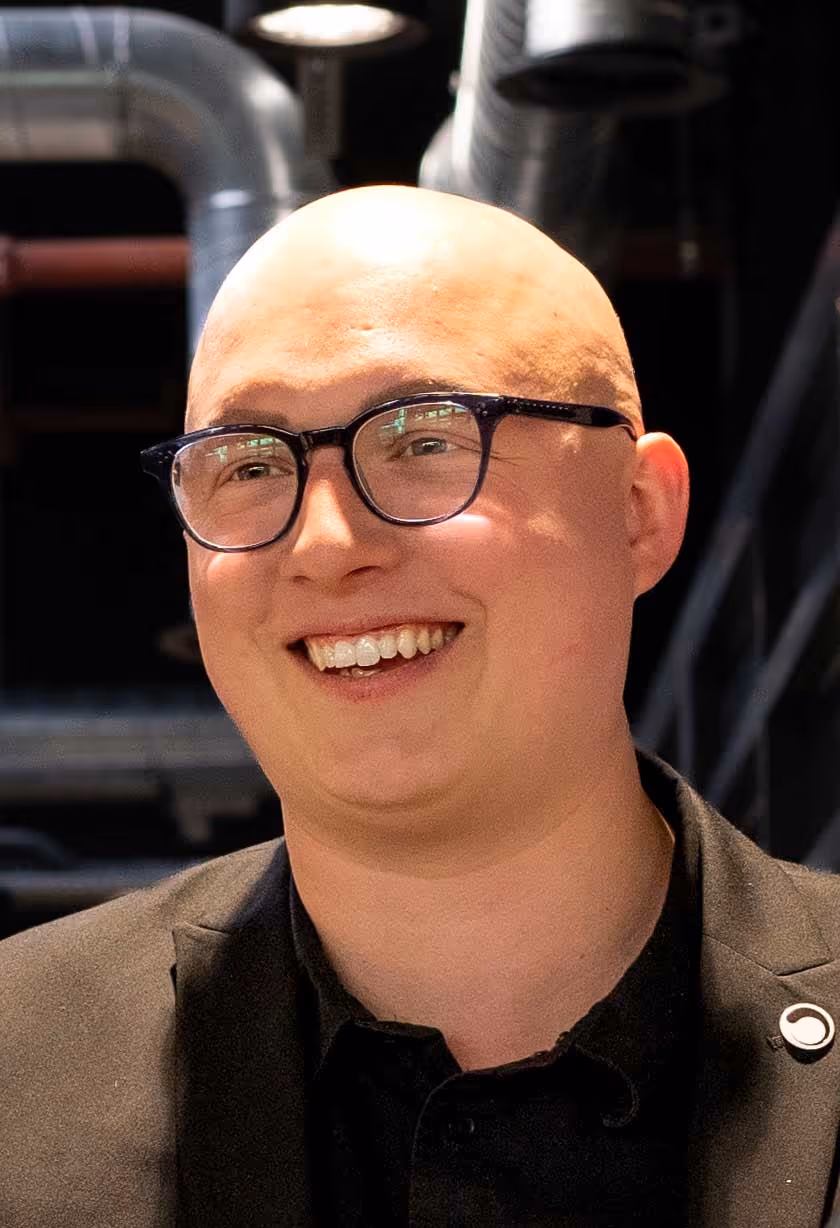A person with glasses smiles brightly while wearing a dark blazer.