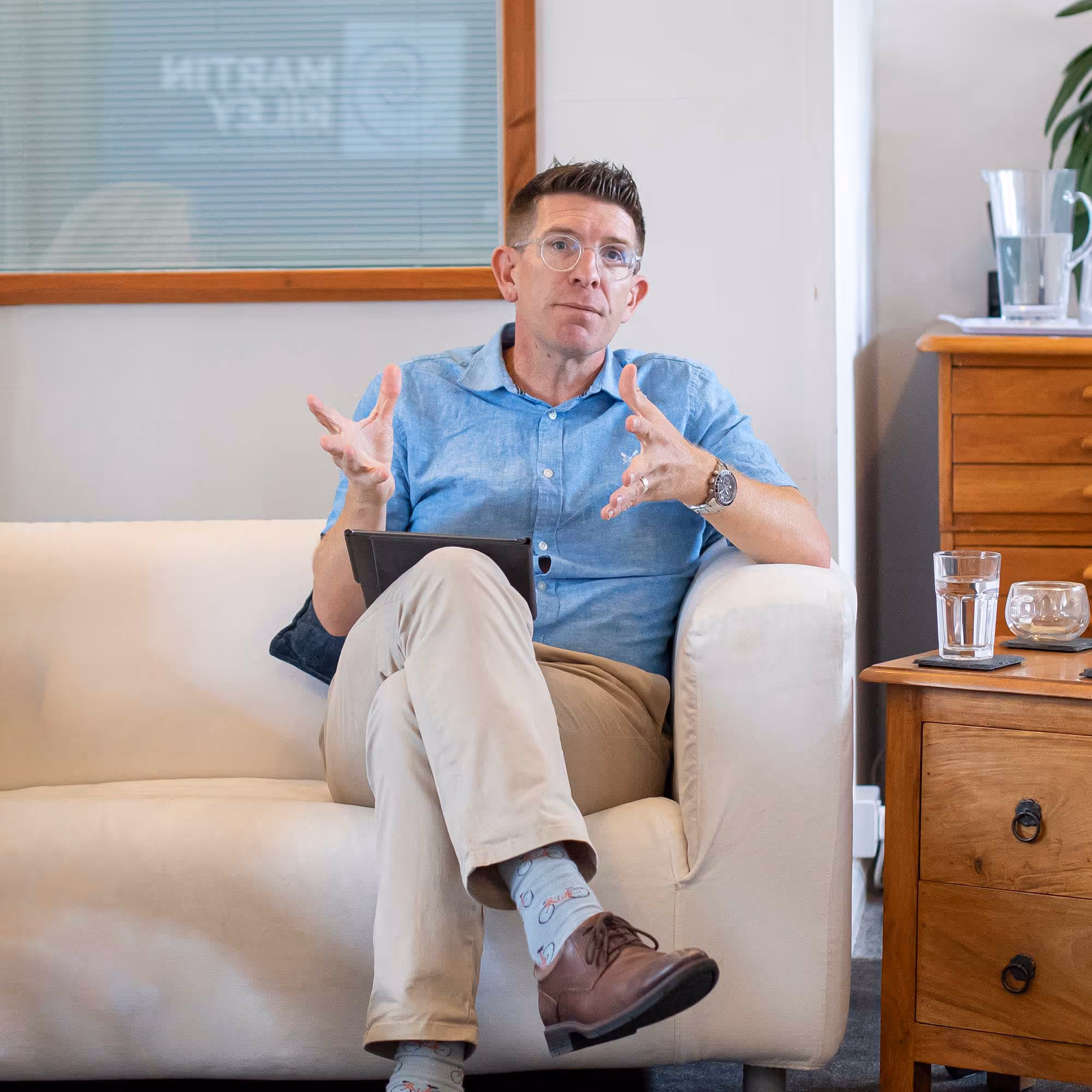 Person sitting on a sofa, gesturing with hands, while speaking and holding a device on their lap.