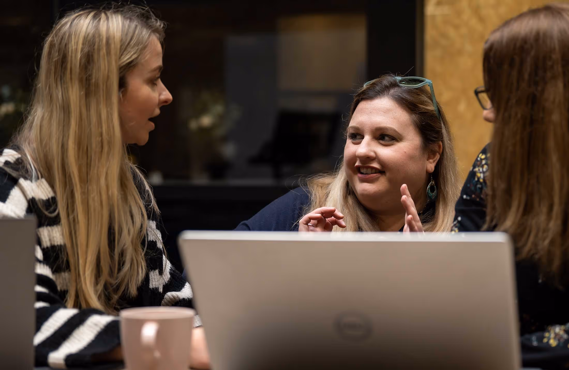 Three women sitting at a table, engaged in conversation with laptops open in front of them.