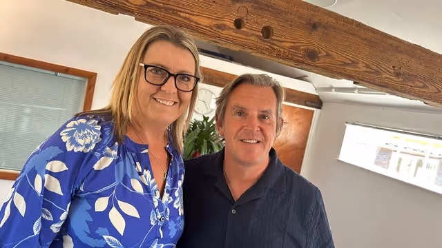 Two people smiling indoors, in front of a wooden beam.