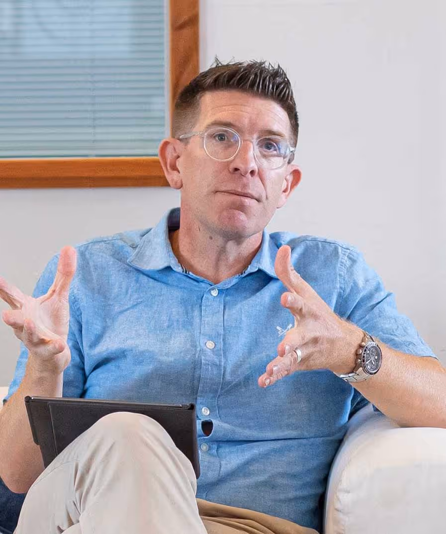 Man in blue shirt sitting on a couch, gesturing with hands, holding a digital tablet.