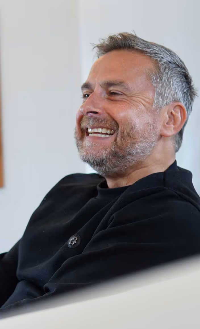 Man with gray hair and beard smiling while sitting indoors, wearing a black sweater.