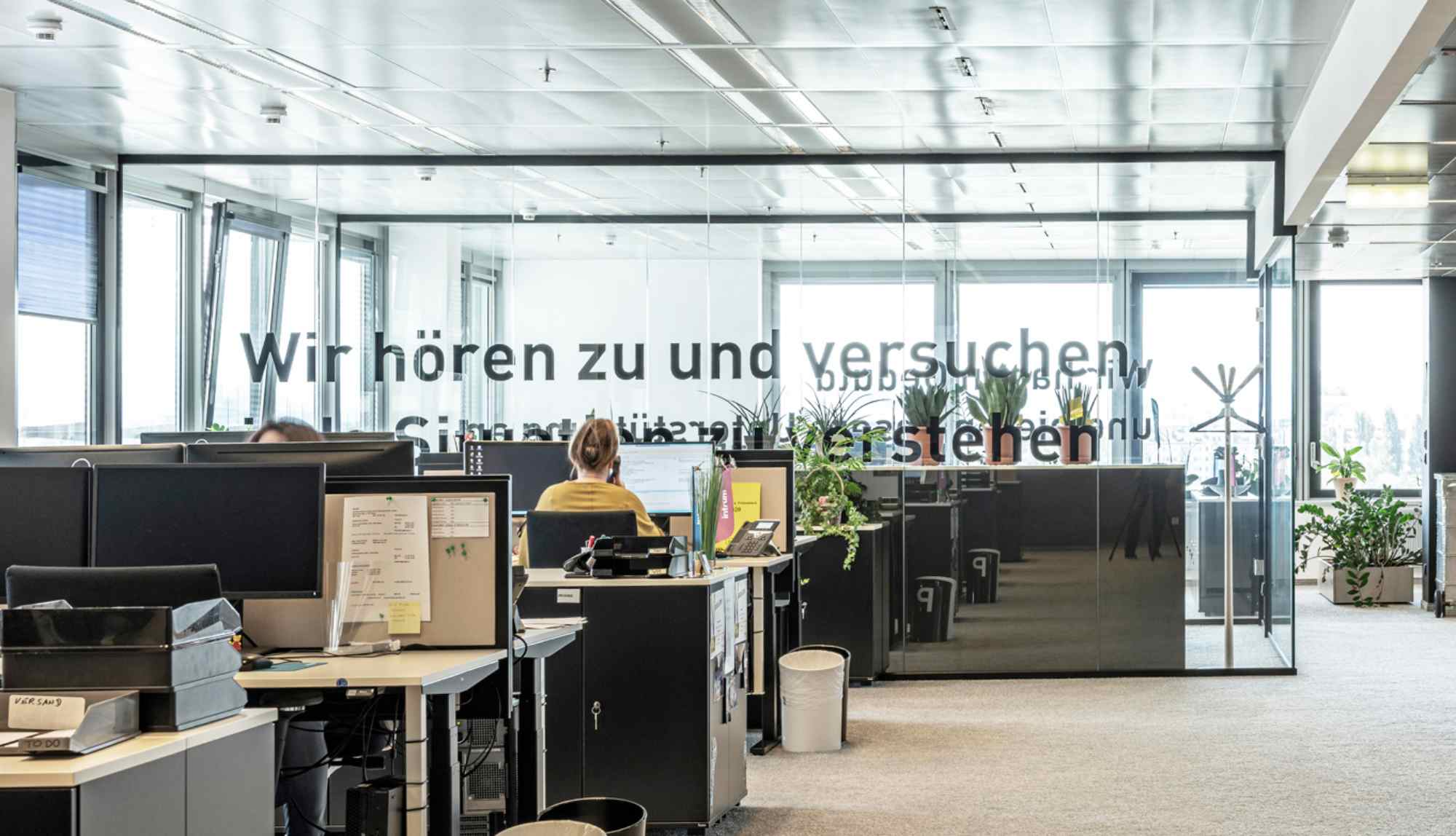 Großraumbüro mit Glasfront und Text „Wir hören zu und versuchen zu verstehen“