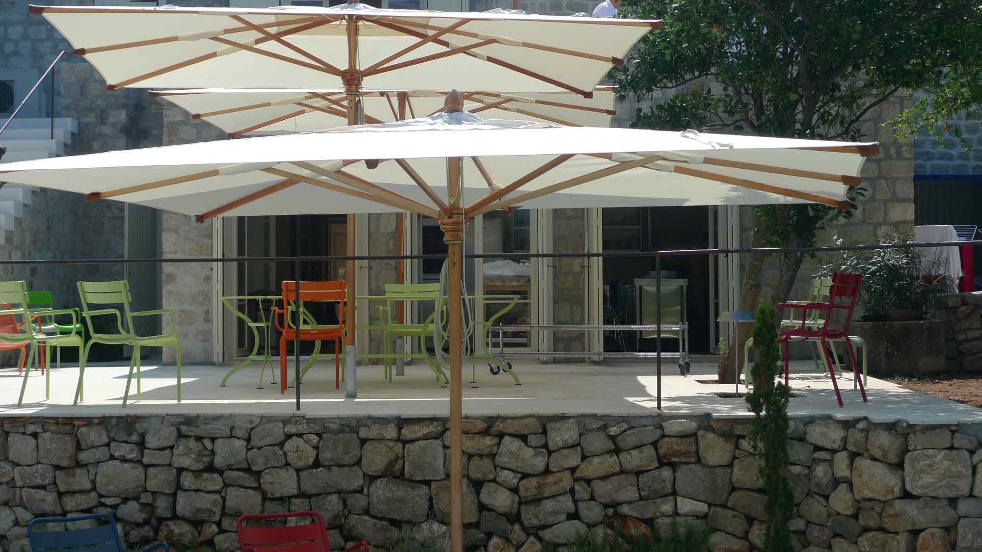 Essbereich mit bunten Stühlen auf Terrasse über Natursteinmauer, geschützt durch Sonnenschirme.