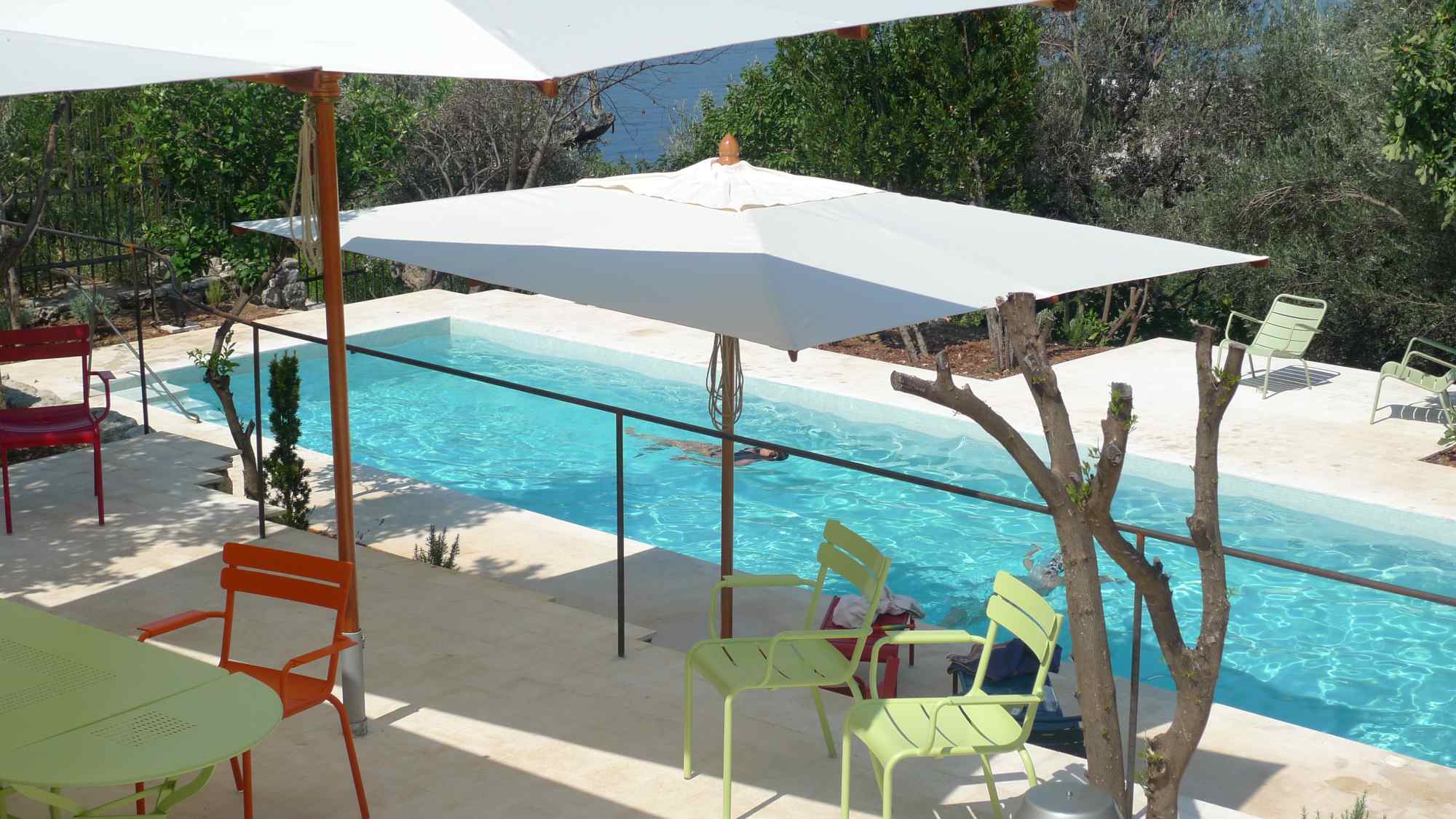 Sitzgruppe mit bunten Stühlen unter weißen Sonnenschirmen direkt neben Pool und Olivenbaum.