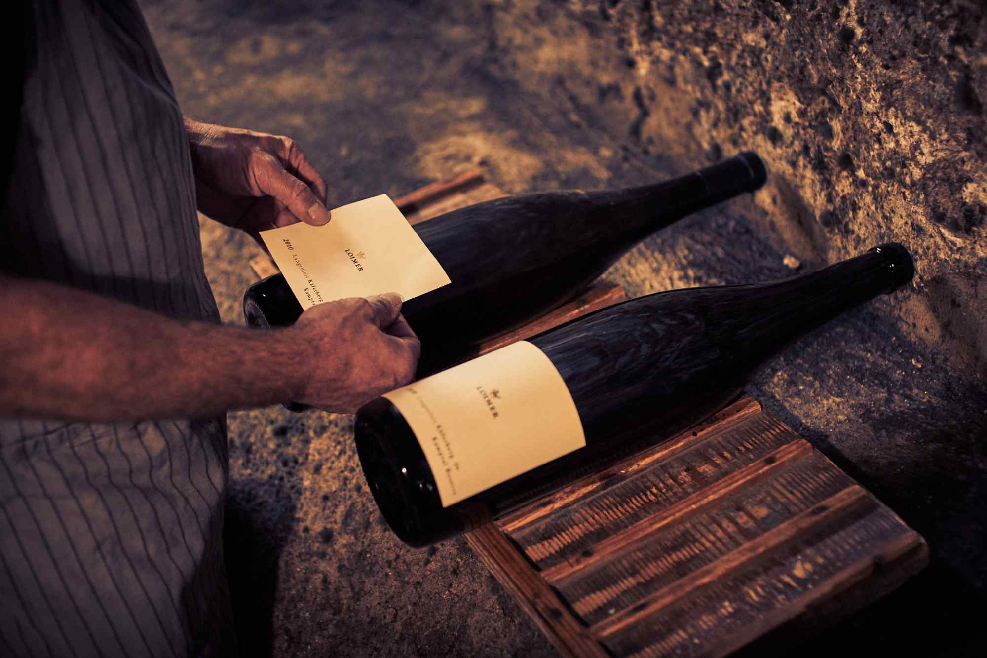 Zwei Hände sortieren mehrere Loimer-Weinflaschen in einem Weinkellerregal.