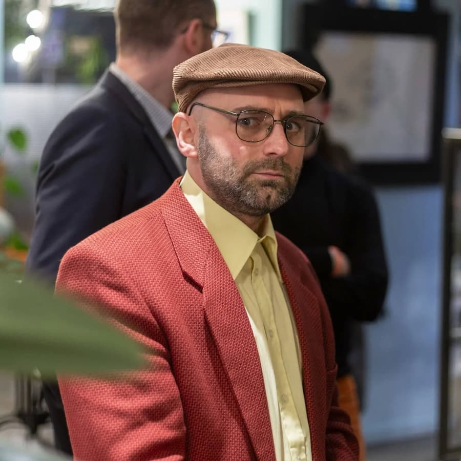 un homme à lunettes fumées, béret sur la tête, chemise jaune et veste rouge