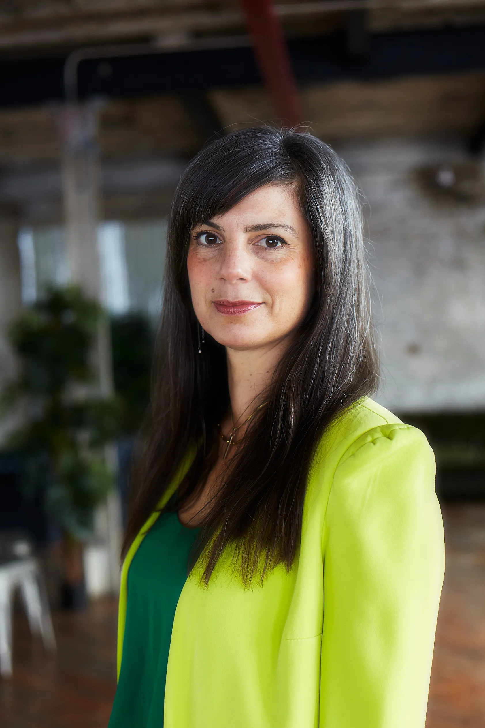 Woman with long dark hair wearing a bright yellow blazer and green top, standing indoors with a blurred background.