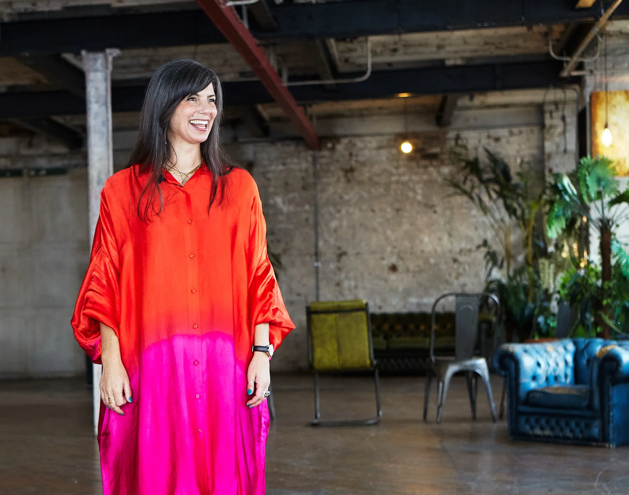 Smiling woman with long dark hair wearing a bright orange and pink dress standing in a spacious room with industrial decor and chairs.