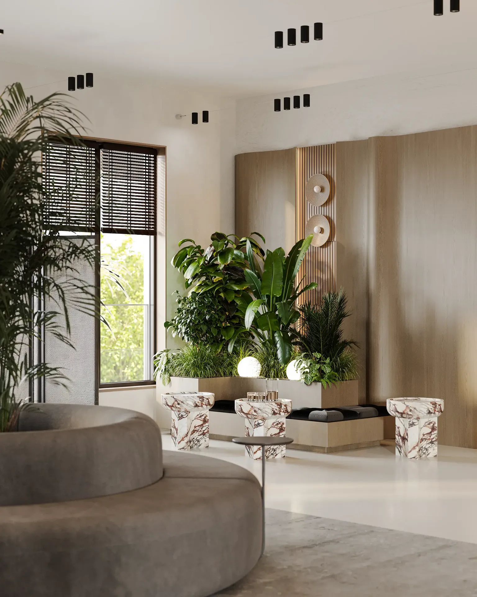 Modern lounge area with curved gray sofa, marble stools, lush indoor plants, and natural light from large window with blinds.