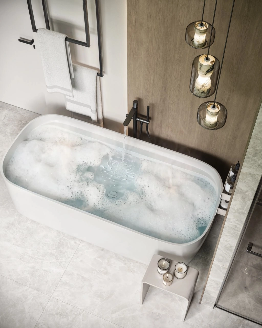 Modern white bathtub filled with bubble bath water, black faucet running, with candles on nearby stool and towels hanging on black rack.