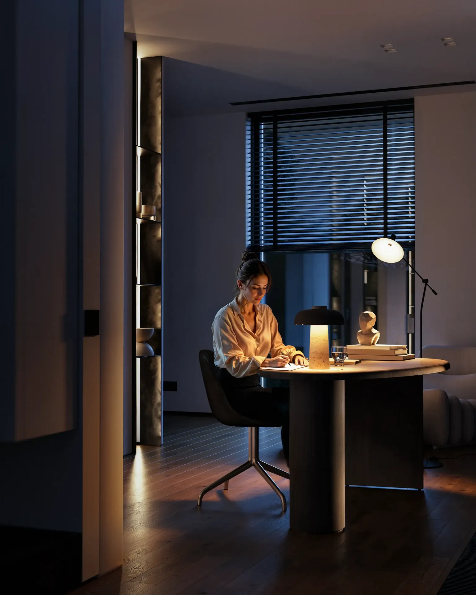 Cozy evening view of a home office, showing a woman writing at a desk under warm ambient light.