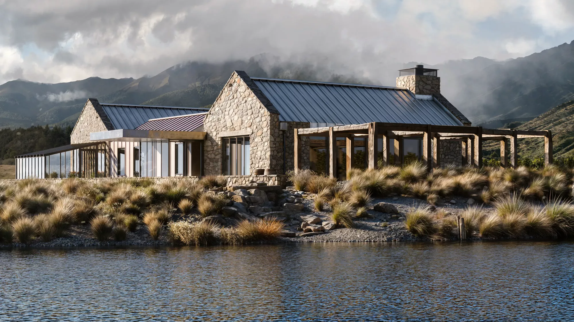 A rustic-modern stone lake house with a standing-seam metal roof and wooden pergola, situated on a bank of tussock grass overlooking a lake, with mist-covered mountains in the background