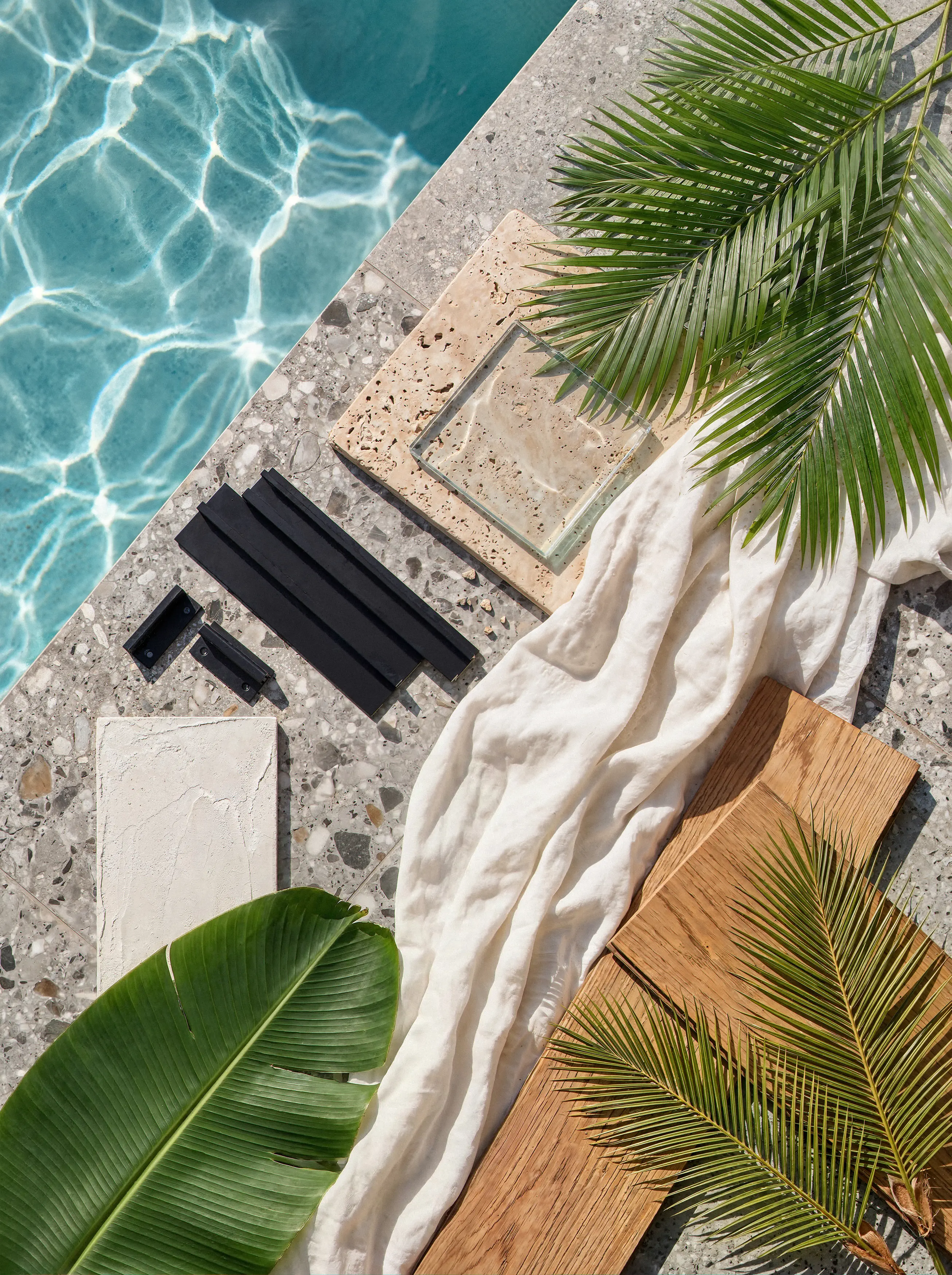 Architectural material mood board flat lay featuring terrazzo, warm wood planks, travertine stone, black metal, and white linen beside a sunlit swimming pool.