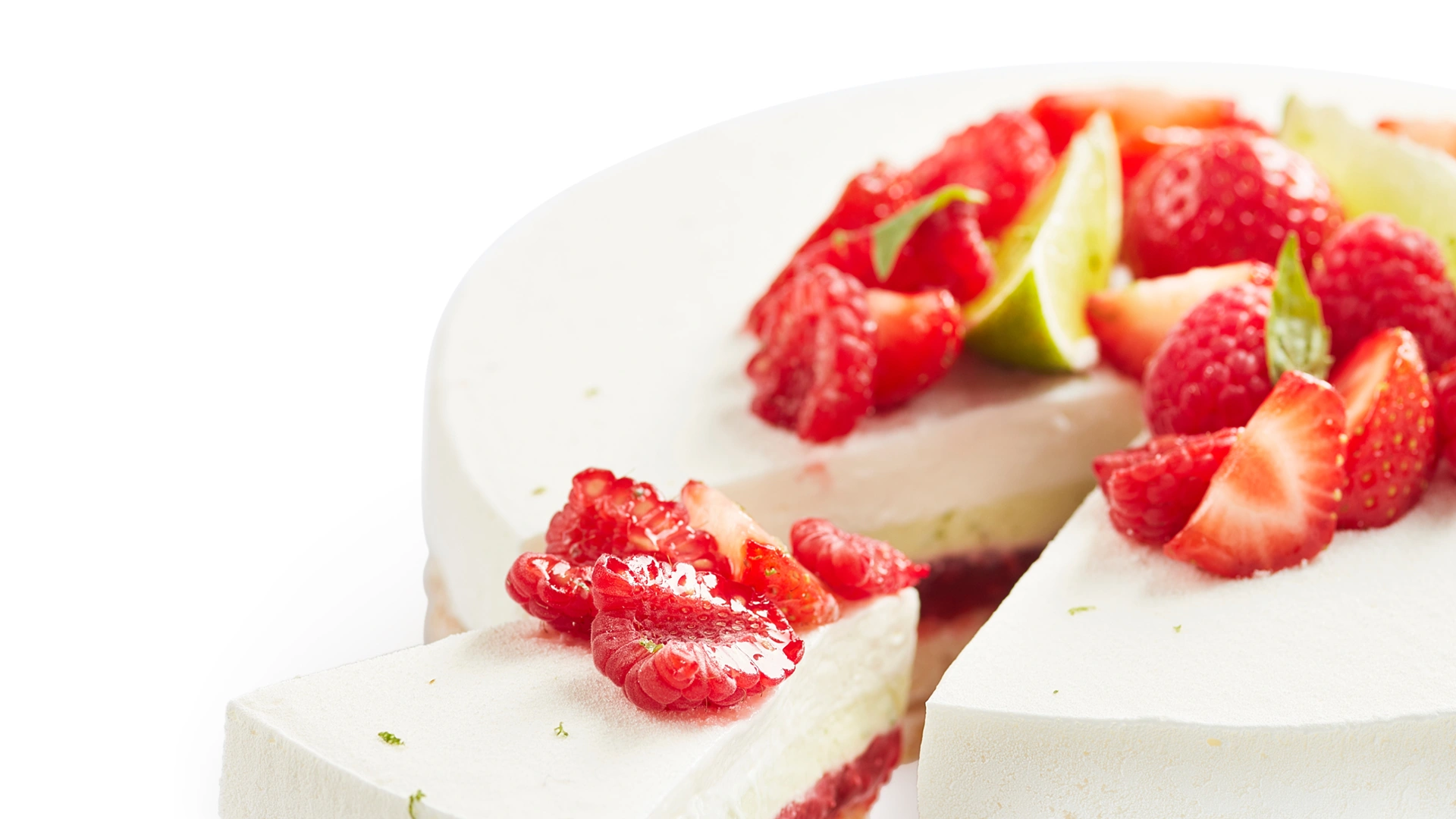 A slice of a layered mousse cake with a crispy puffed rice base, a middle layer of red fruit jelly, and a smooth white mousse topping, garnished with fresh raspberries and lime slices.