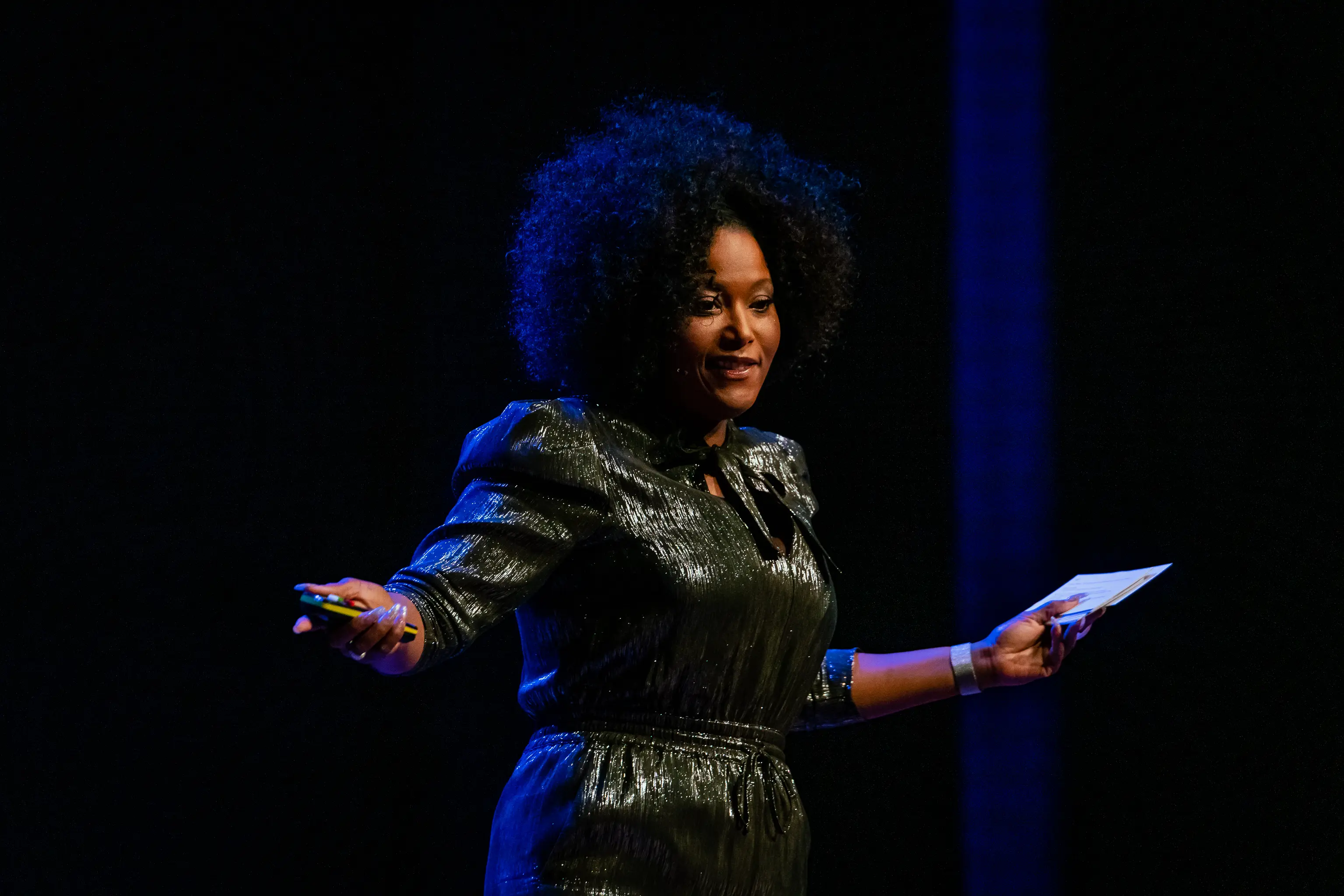 Denice Dest staat in een kleurrijke outfit voor een groep deelnemers en begeleidt hen actief tijdens een interactieve sessie.