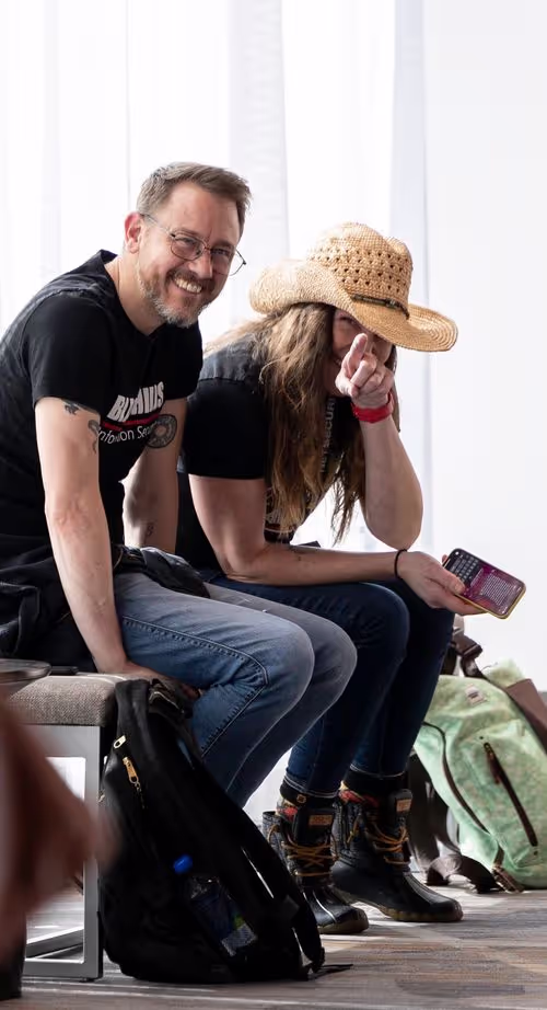 Man and woman sitting down, smiling and looking at the camera, woman is pointing at the camera