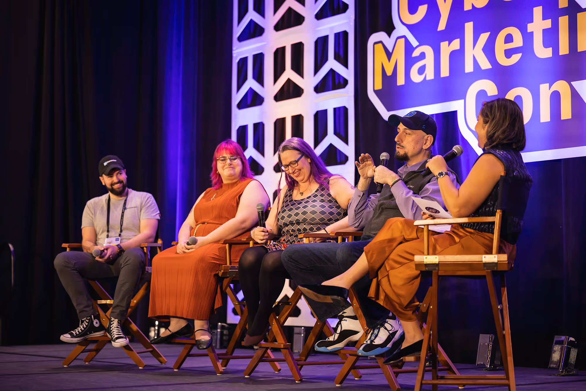 Five speakers sitting on chairs on the stage behind a poster of cyber marketing con
