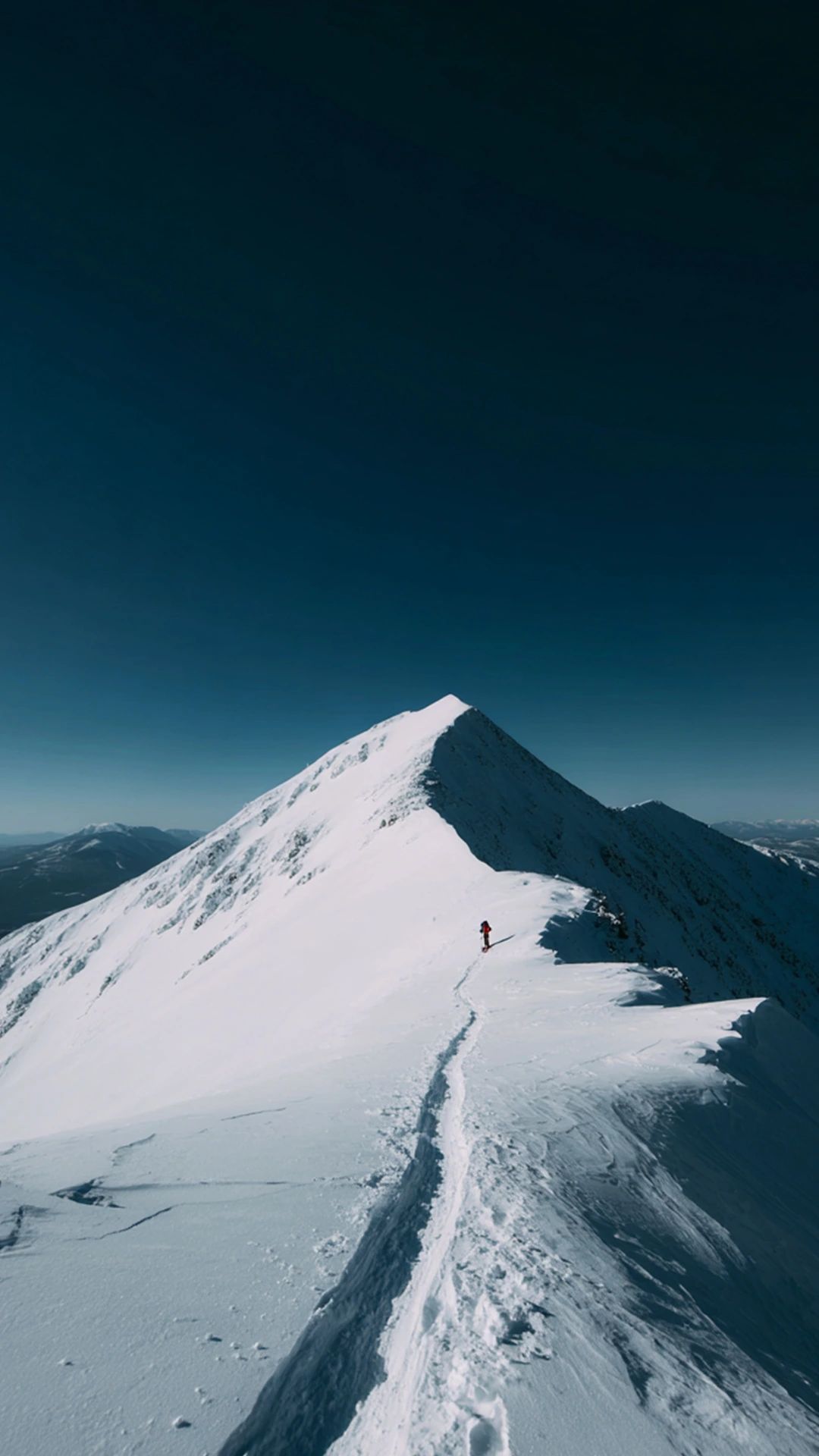 A person walking on a mountain top, filled with snow. The right side is covered in shadow, the left side is in the sun. In the distance, there are other mountain tops.