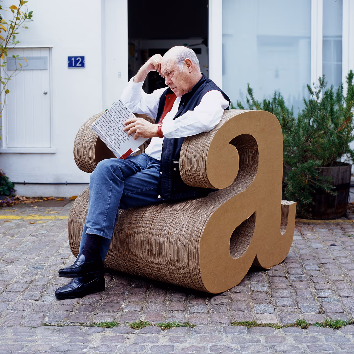 Alan sitting on a cardboard chair, shaped into a lowercase letter a, whilst reading his book The art of looking sideways.