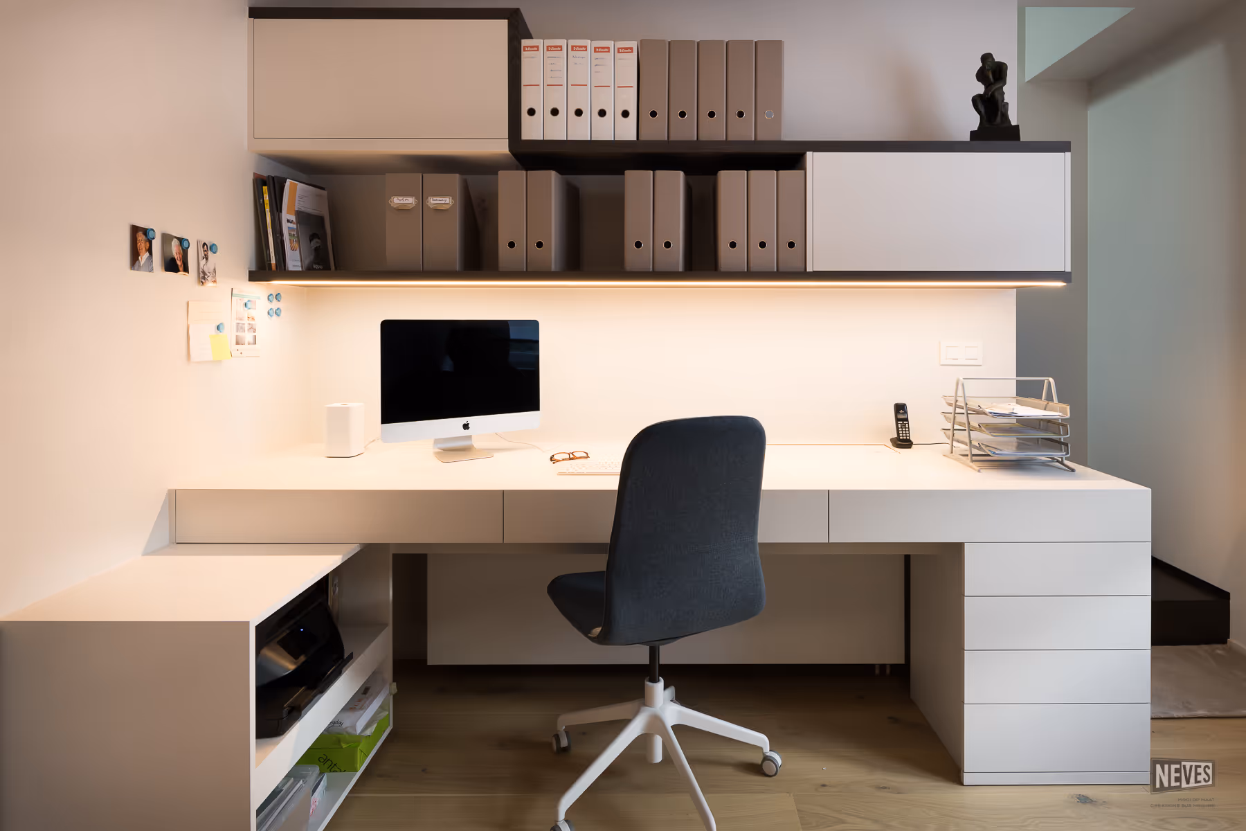 Minimalistisch wit bureau met zwarte bureaustoel, computer, ordners aan de wand en een bureaulamp eronder.