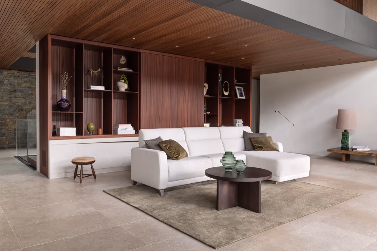 Modern woonkamer met een wit hoekzetel, een ronde houten salontafel met groene vazen, een bruine kast met decoraties en een houten plank met lamp en boeken.