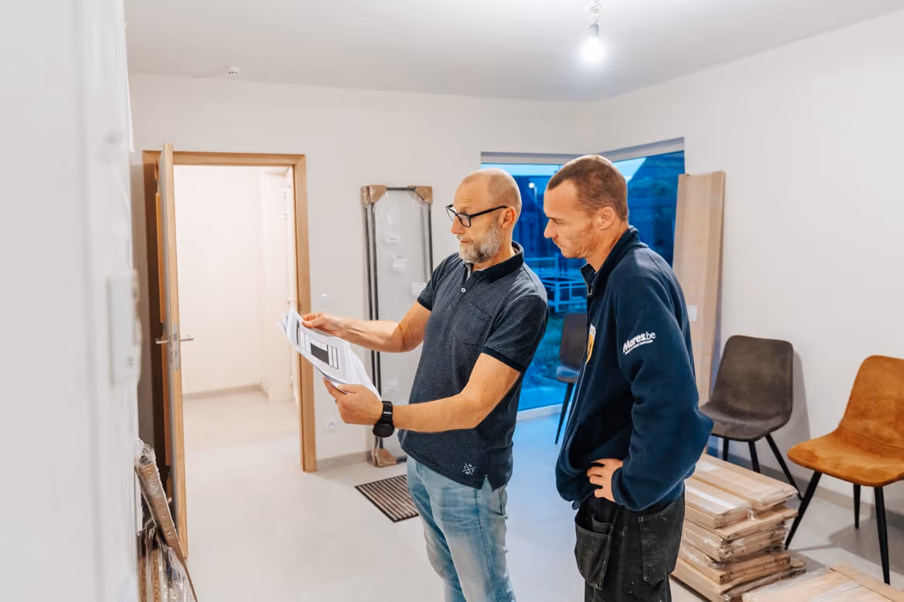Twee mannen bespreken plannen en kijken naar documenten in een kamer met verhuisdozen en stoelen.