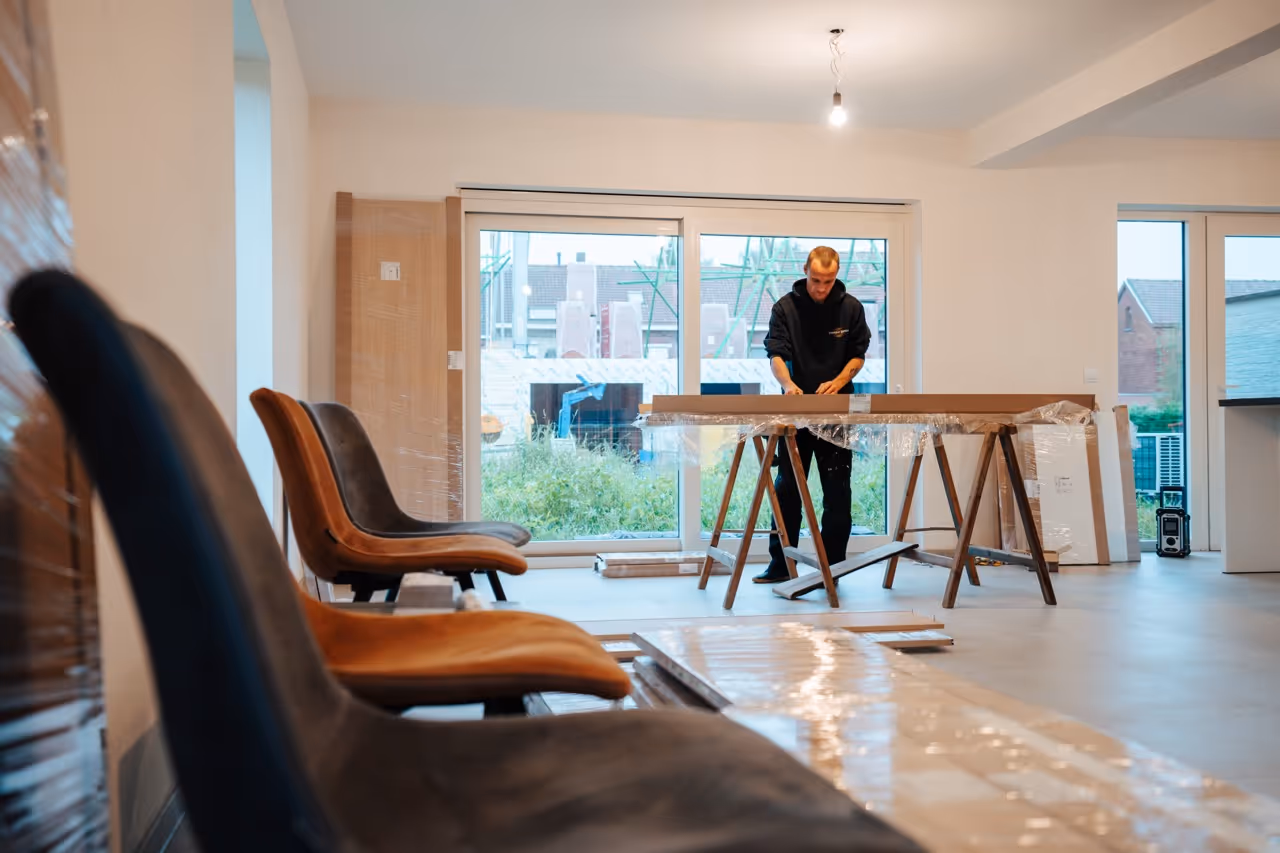 Man werkt aan een houten plank op een steigerconstructie in een lege kamer met grote ramen en stoelen op de voorgrond.