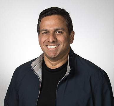 Smiling man with dark hair wearing a black shirt and navy jacket against white background.