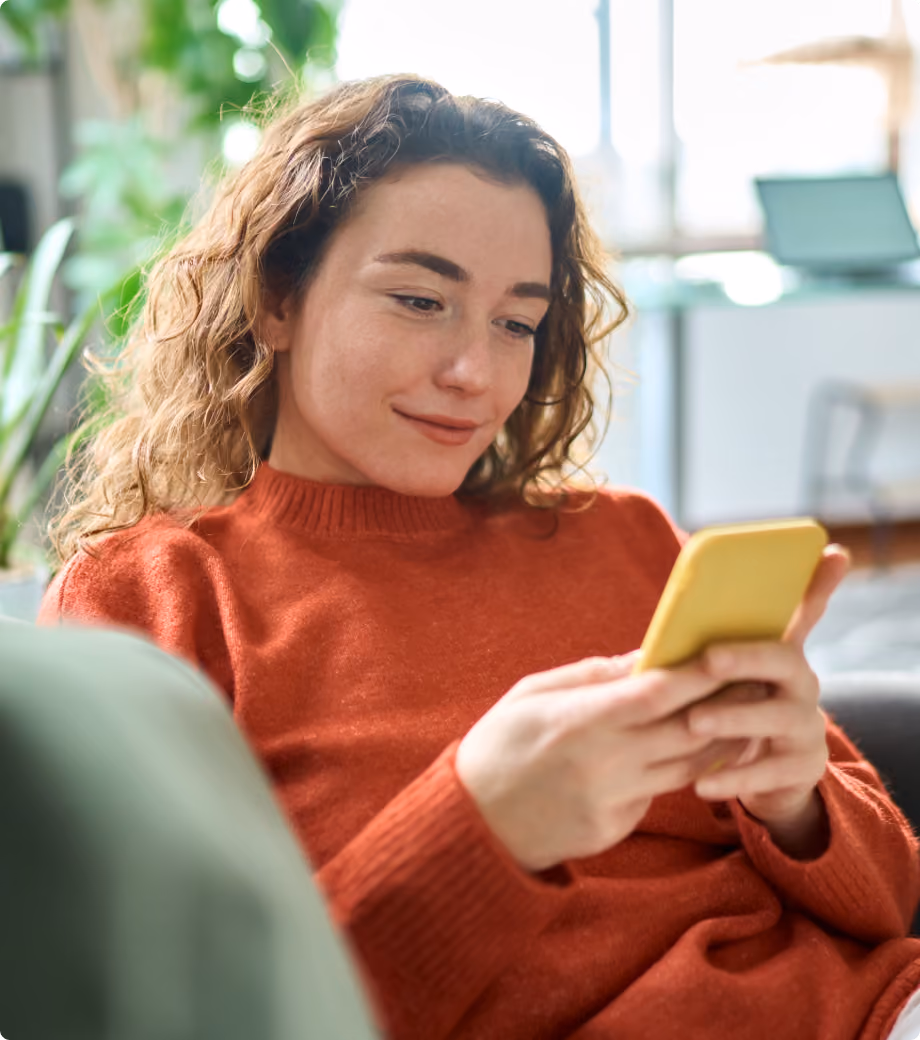 Woman in orange sweater looking at a yellow smartphone while sitting indoors on a couch.