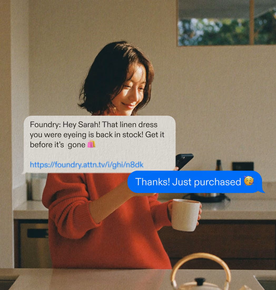 Woman in red sweater holding a mug and smartphone, smiling at a text message from Foundry about a dress restock.