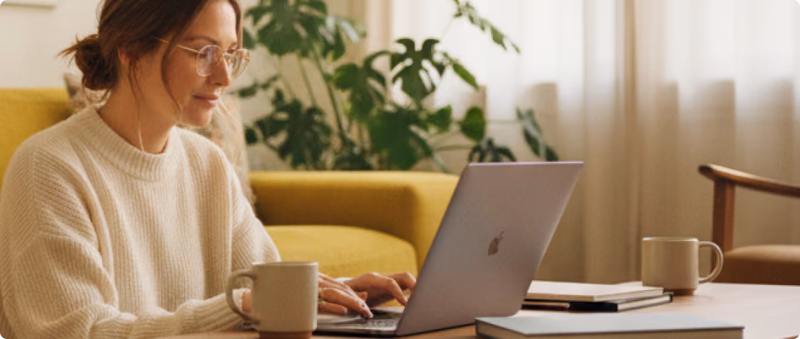 Woman wearing glasses and a cream sweater typing on a laptop with a coffee mug on the table.