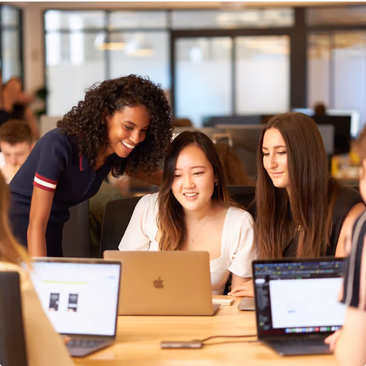 Three women collaborating around a laptop in a modern office setting, smiling and focused.