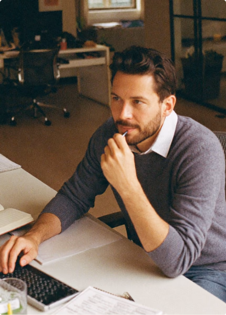 Man with beard and sweater seated at desk, working on a computer while holding a pen near his mouth.