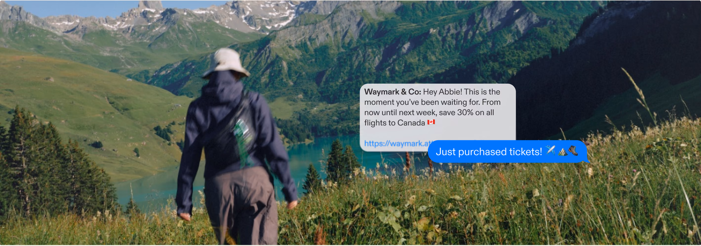 Person wearing a hat hiking towards a lake in a mountain valley under a blue sky.