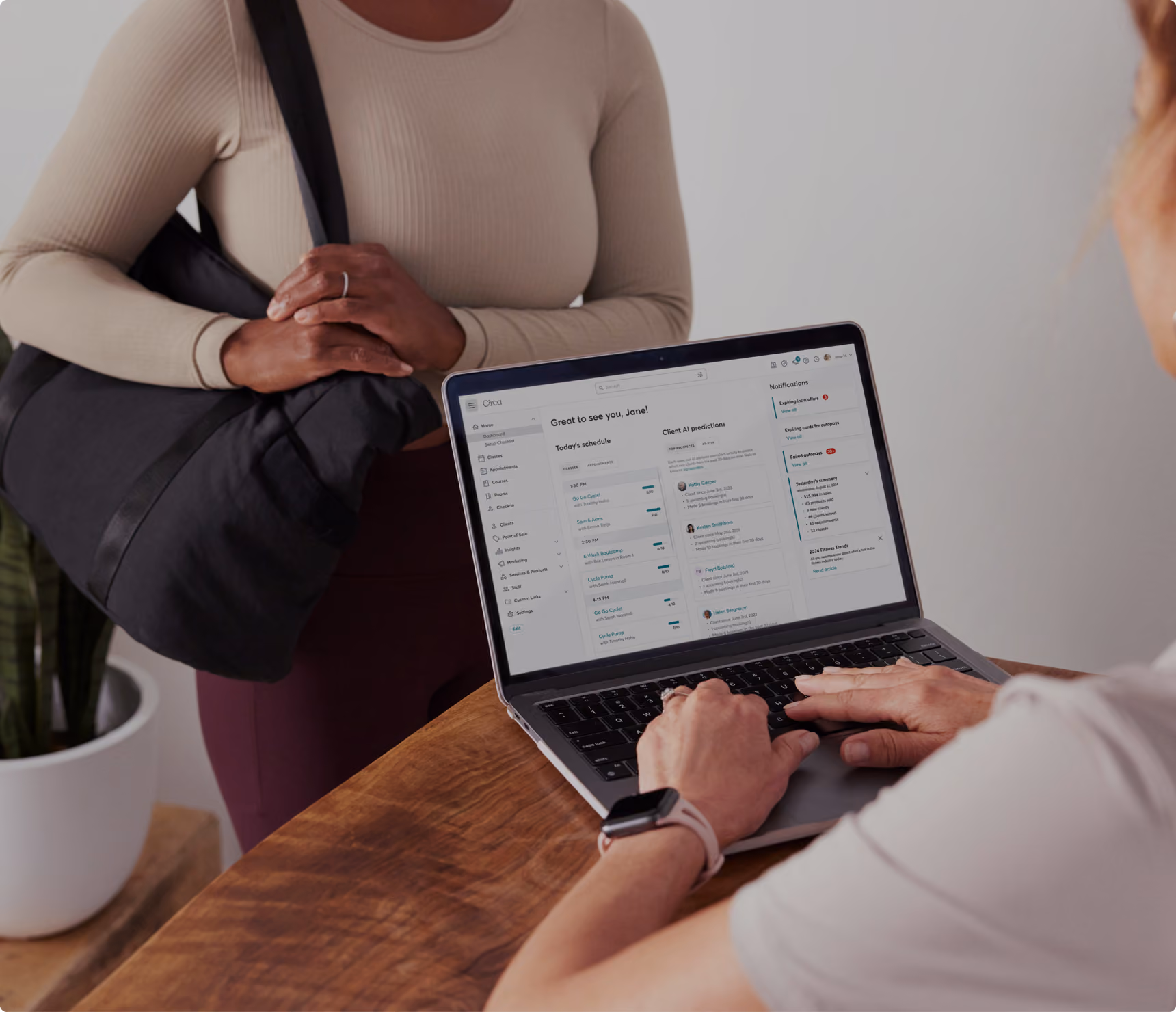 Person typing on a laptop showing a client scheduling and notifications dashboard, another person stands nearby.