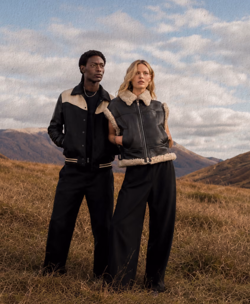 Man and woman wearing black shearling jackets standing in a grassy field with mountains in background.