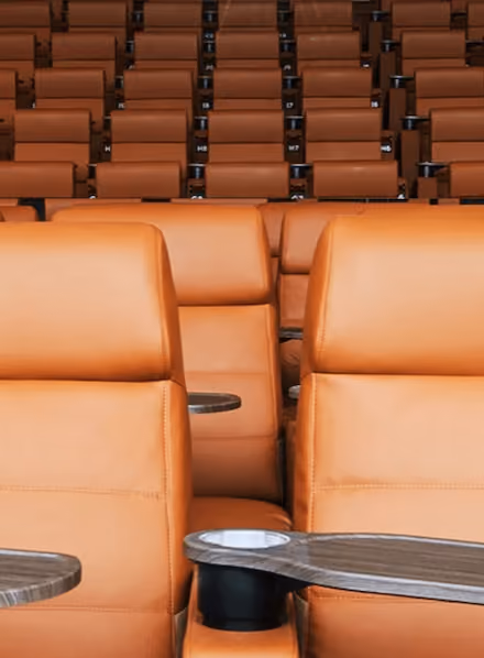 Rows of empty tan leather recliner seats with attached wooden trays in a theater.