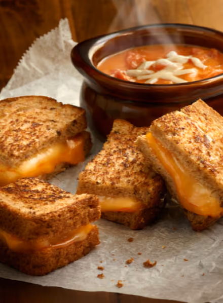 Four grilled cheese sandwich quarters on parchment paper with a bowl of tomato soup topped with shredded cheese.